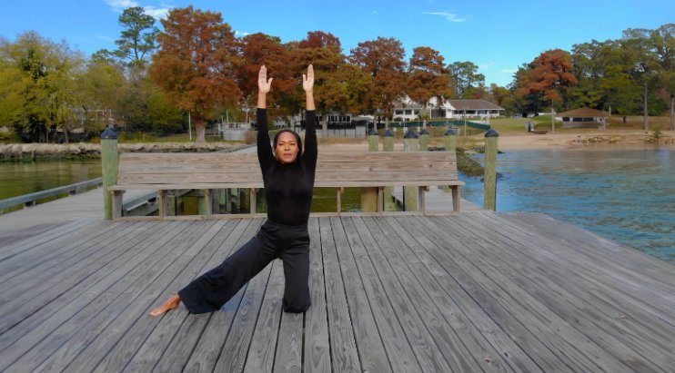Carmen Kennedy in a wide kneeling pose with arms raised on the dock, autumn trees across the water